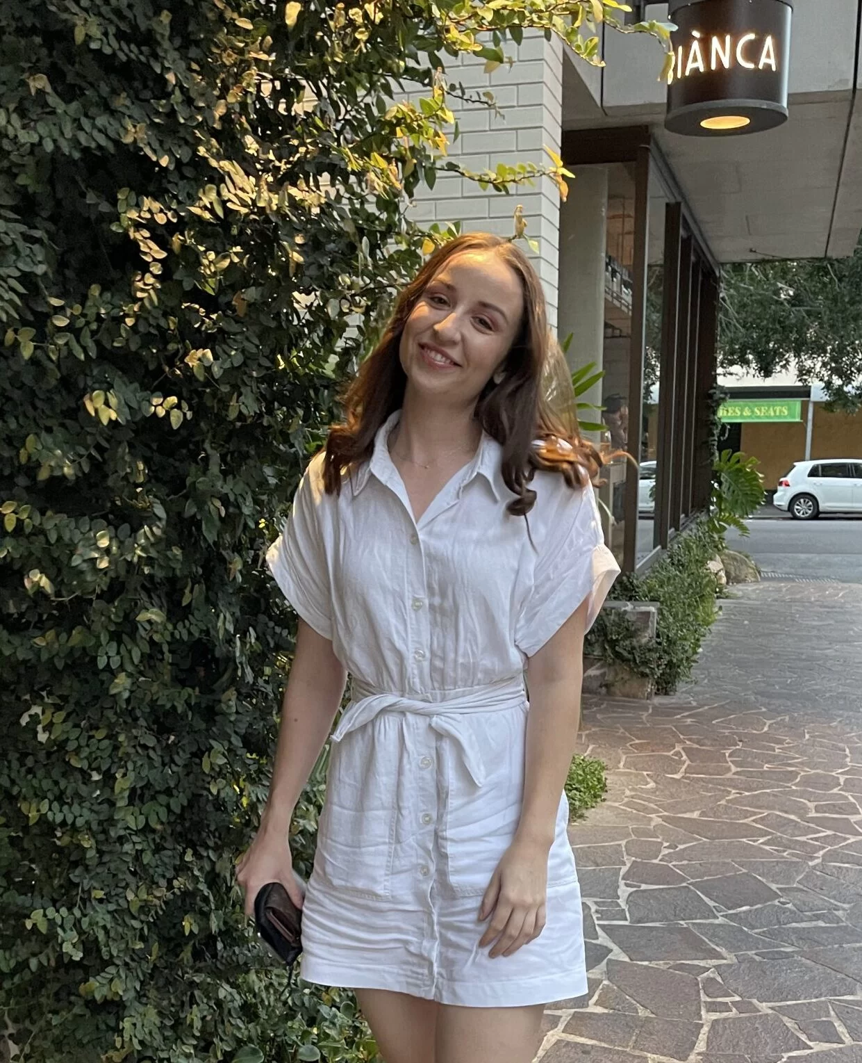 A girl in a white dress stands smiling in front of some bushes